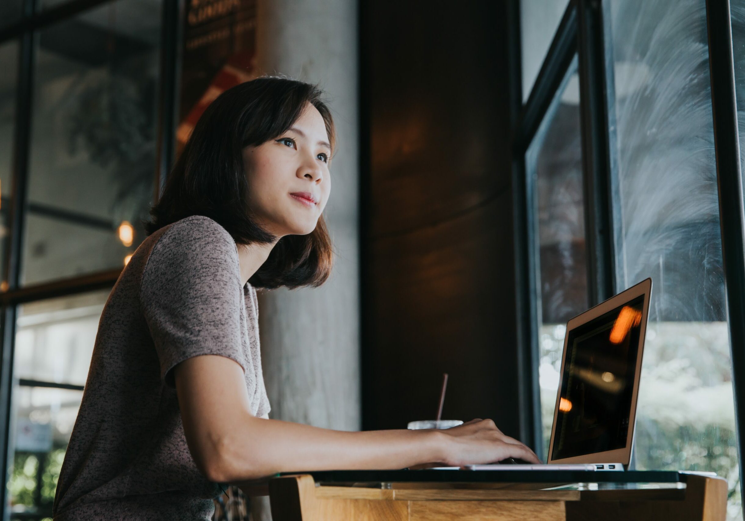 Beautiful young Asian woman working at a coffee shop with a laptop. Female freelancer connecting to internet via computer. Casual Thinking Browsing Concept.